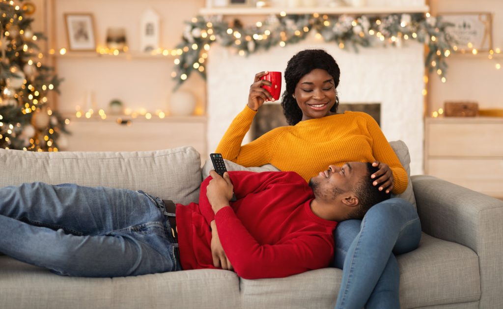 Relax at home on couch and New Years mood. Millennial smiling african american guy lies on knees of lady with remote control, lady holds cup with hot drink in interior with Xmas tree with garlands