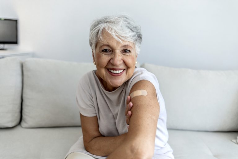 Portrait of a senior woman proudly showing her arm with bandage after getting vaccine. Mature white haired woman sitting against bright background after receiving coronavirus vaccination, thumbs up.