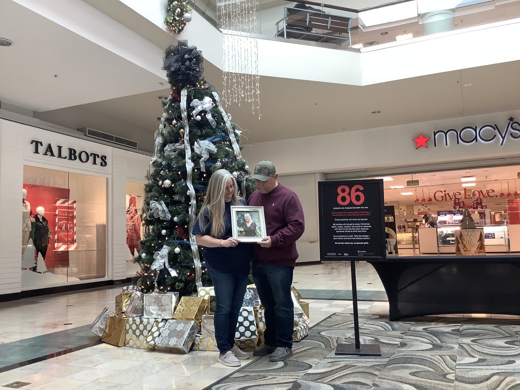A woman and man stand in front of a decorated Christmas tree at Woodland Hills Mall. The woman is holding a picture of a loved one who was killed by a drunk driver.