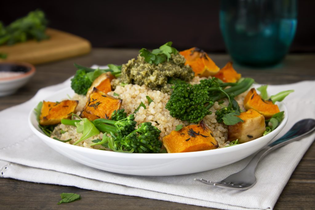 A Quinoa bowl with roasted butternut squash, broccolini, leafy greens and pesto. Shot on a dark wooden table with fresh vegetables and Himalayan salt in the background. Healthy vegetarian food recipe.