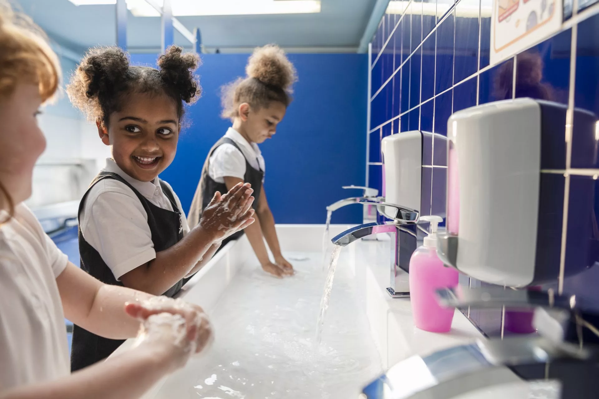 A side-view of diverse young schoolgirls washing their hands with soap and water in their school bathrooms. They are all wearing their school uniforms.
