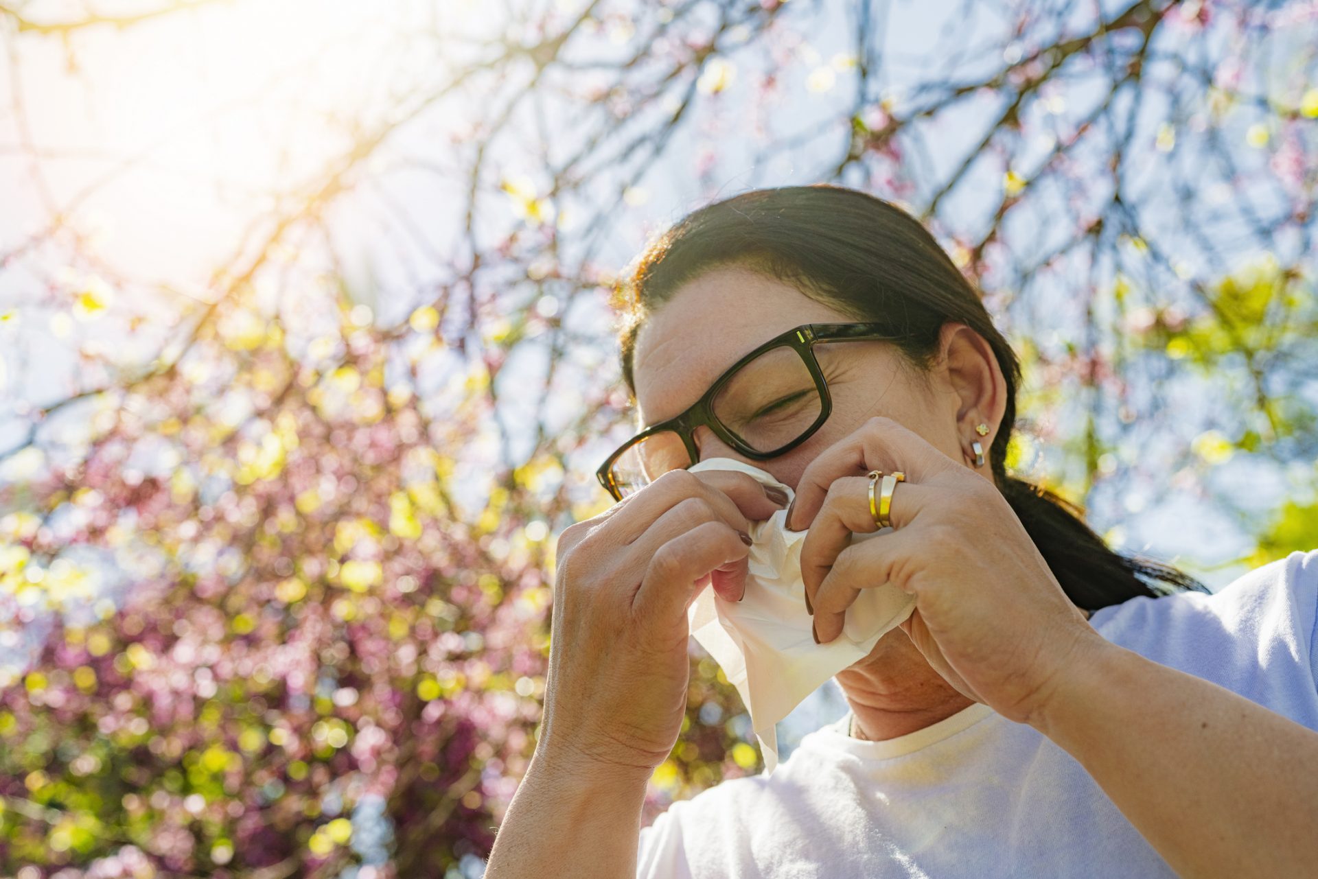 Middle-age woman with seasonal allergy using paper tissue to blow her nose while outside under a blooming tree.