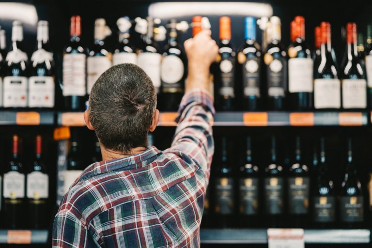 Rear view of a man reaching for a bottle of wine while shopping in the aisle of a liquor store