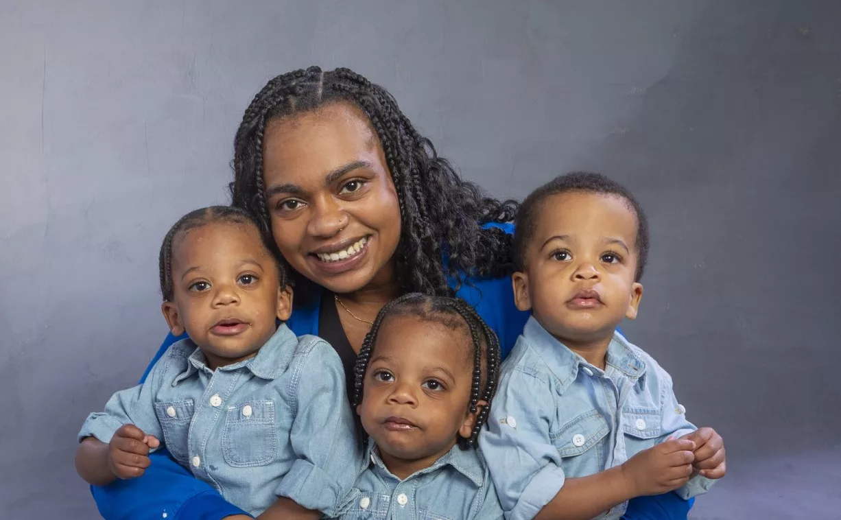 A woman wearing a royal blue blazer sits on the floor smiling while holding three young children in matching outfits.