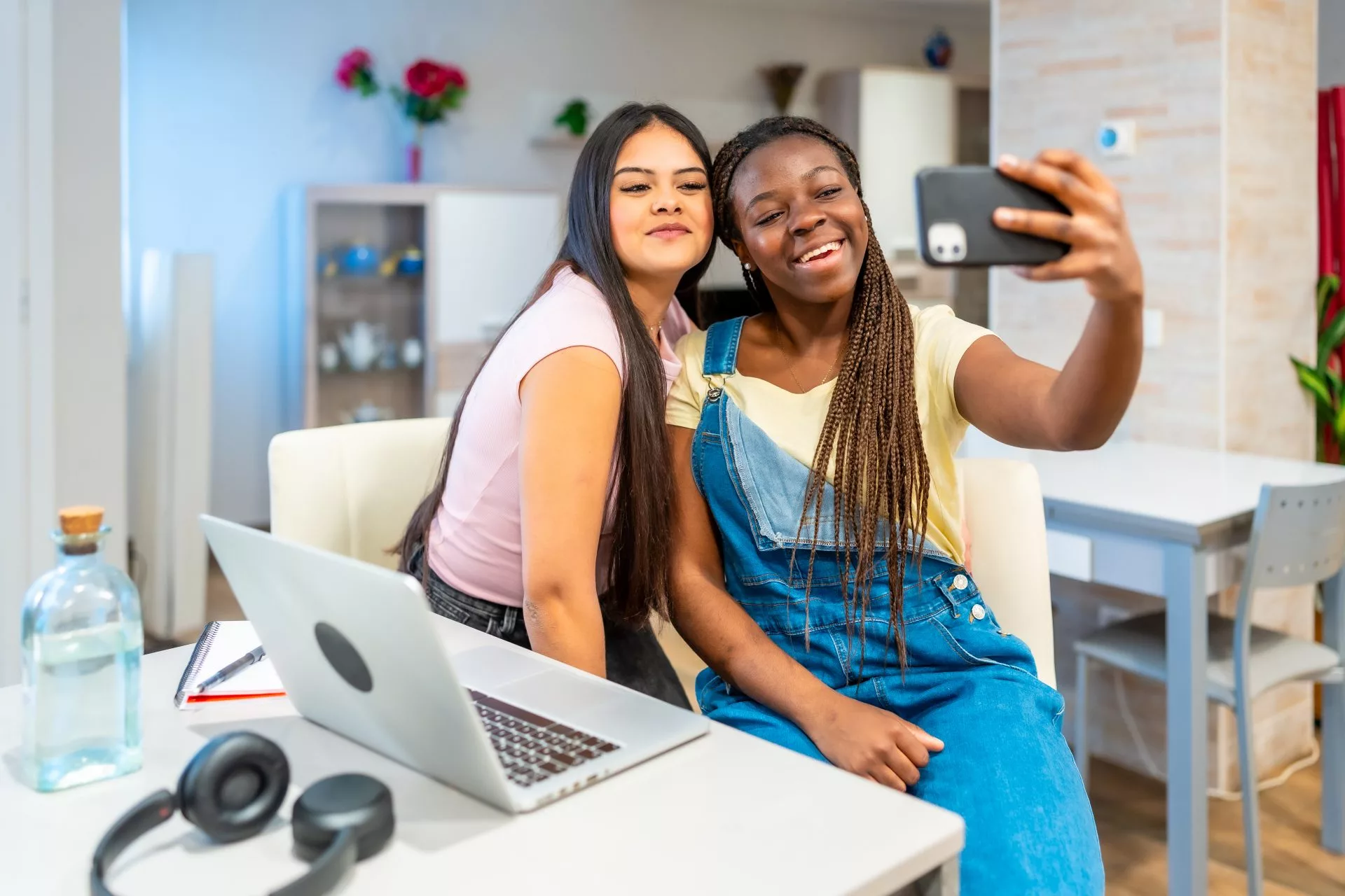 Two young women smile while taking a selfie together at a table with a laptop and notebook.