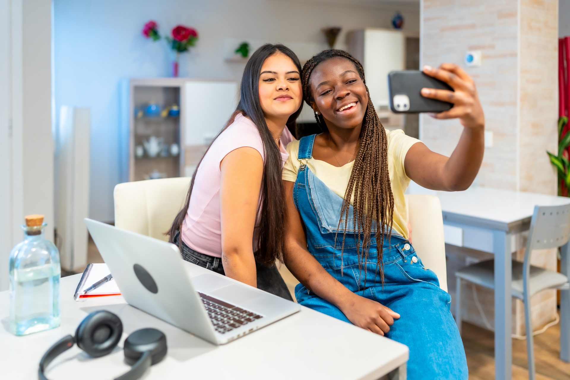 Two young women smile while taking a selfie together at a table with a laptop and notebook.