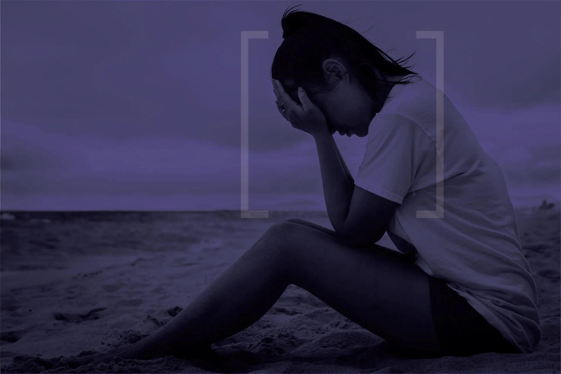 Young woman sitting outdoors (likely on a beach or shoreline) with her head down, holding her face in her hands